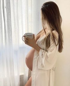 a pregnant woman holding a coffee cup in front of a window