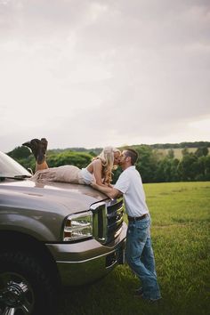 a man and woman kissing on the hood of a truck in a field with trees