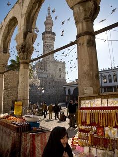 a woman standing in front of an archway with birds flying around and buildings behind her