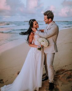 a bride and groom standing on the beach at sunset with their arms around each other