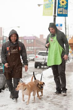 two men walking dogs in the snow on a city street with cars and signs behind them