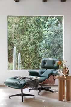 an eames lounge chair and ottoman in front of a large window with trees outside