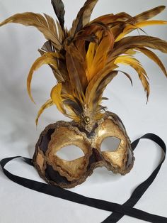 Elegant Venetian mask adorned with lush golden feathers, offering a refined and majestic look. Made according to the Venetian artisanal tradition, this mask is perfect for masked balls, carnivals and costume parties. It features detailed gold hardware and a central embellishment with a gold-tone costume jewel, adding a touch of sophistication to the overall look. Gold Masks And Prosthetics For Carnival, Gold Masks For Carnival, Gold Baroque Masks For Mardi Gras, Carnival Feather Eye Mask, Gold Baroque Masquerade Mask For Carnival, Gold Baroque Mask For Costume Party, Carnival Festival Eye Mask, Vintage Masks And Prosthetics For Mardi Gras Carnival, Feathered Masquerade Mask For Costume