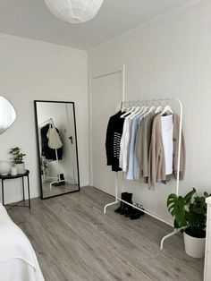 a bedroom with white walls and wooden flooring, clothes on hangers, mirror and potted plant