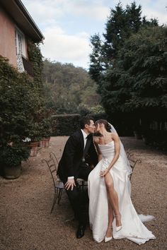 a bride and groom kissing on a bench in front of a house with trees behind them