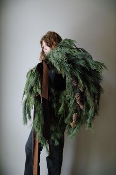 a woman is holding a wreath made out of pine cones and evergreen needles, while standing against a white wall