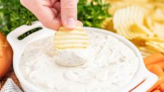 a hand dipping a cracker into a bowl of dip surrounded by carrots and celery