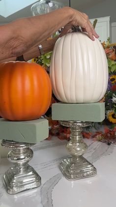 two pumpkins sitting on top of pedestals in front of a flower arrangement and a woman's hand reaching for one