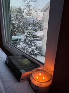 a candle and book on a bed in front of a window with snowy trees outside