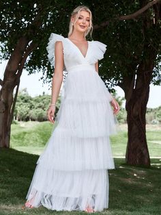 a woman standing in front of trees wearing a white dress with ruffles on the skirt