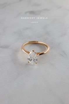 a gold engagement ring with a pear shaped diamond on the front and side, sitting on a marble surface