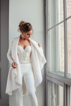 a woman standing next to a window wearing white pants and a jacket with lace on it