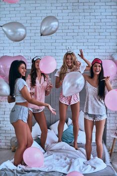 three women in pajamas and headbands are jumping on a bed with pink balloons