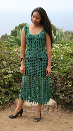 a woman wearing a green crochet dress standing in front of bushes and plants