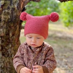 a small child wearing a knitted hat with pom - poms standing in front of a tree