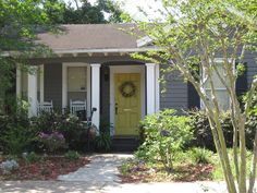 a yellow door sits in front of a gray house with trees and bushes around it