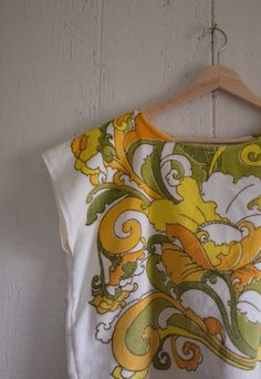 a white shirt with yellow and green designs hanging on a wooden hanger next to a wall