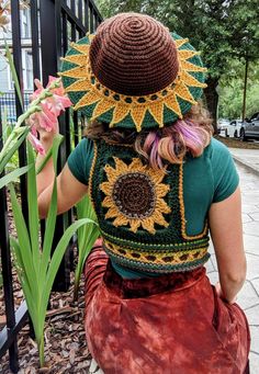 a woman wearing a crocheted sunflower hat sitting on the ground next to a fence