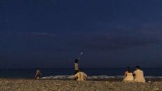 three people sitting on the beach flying a kite in the sky at night with other people nearby