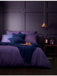 a bed with purple sheets and pillows in a room next to a lamp on a table