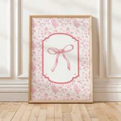 an empty frame with a pink bow on it in front of a white wall and wooden floor