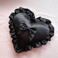 a black heart shaped pillow with a bow on the top and bottom, sitting on a pink surface