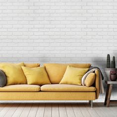 a yellow couch with pillows on it in front of a white brick wall and wooden floor