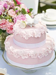 a three tiered pink cake with white flowers on the top and bottom is sitting on a glass platter