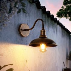 an outdoor light on the side of a building next to a wall with flowers and trees