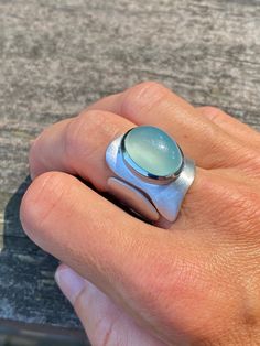 This one is a true eye catcher. Very modern and contemporary adjustable sterling silver ring with perfect diamond aqua chalcedony cabochon. Looks amazing and very hip on when worn. Asymmetrical shape of ring will surely make a statement. The wide band is of free -form adjustable design, very original, yet simple. The band is not only has a free - form wavy contour but also slightly etched to give the band a matte finish to match the chalcedony. Very beautiful, industrial and comfortable to wear Modern Adjustable Oval Moonstone Ring, Modern Oval Cabochon Moonstone Ring, Modern Sterling Silver Oval Cabochon Ring, Modern Sterling Silver Ring With Oval Cabochon, Modern Sterling Silver Rings With Oval Cabochon, Modern Oval Cabochon Jewelry With Large Stone, Modern Moonstone Ring With Large Stone, Modern Oval Moonstone Ring With Large Stone, Modern Jewelry With Large Oval Stone