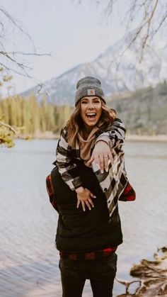 a woman standing in front of a body of water wearing a plaid scarf and hat