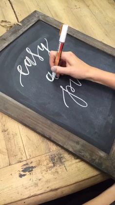 a person writing on a chalkboard with a marker and pen in their hand, while sitting at a wooden table