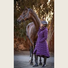 a woman standing next to a brown horse wearing a purple coat and black boots, with trees in the background