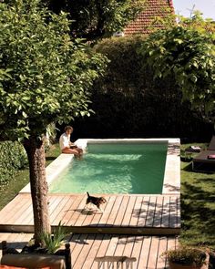 a woman sitting on the edge of a swimming pool next to a tree and chair