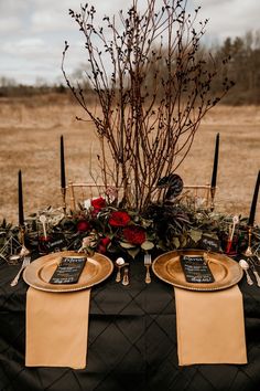 the table is set with black and gold plates, silverware, red flowers and candles