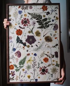 a person holding up a framed painting with flowers and plants on it's side