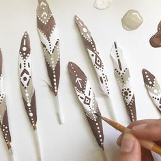 a person is painting some feathers with brown and white designs on them, while another hand holds a pencil