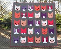 a quilt made to look like cats with glasses on their heads and tails, is displayed in front of a chain link fence