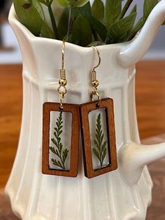 a pair of wooden earrings with green leaves on them sitting in front of a white vase