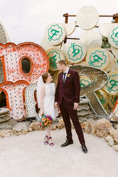 This shot is bursting with charm! 👰🤵 The bride rocks a simple wedding dress, flawless with high neckline elegance, standing beside her groom in a sharp burgundy suit. They're surrounded by retro neon signs that create an eclectic backdrop. Her multicolored bouquet and quirky wedding shoes add a pop of fun! Love the wildflower wedding theme vibe? 🌸 Get inspired here! #weddingideas #unique weddingideas #simpleweddingdress #wildflowerweddingtheme #neonmuseum Wedding Dresses Vegas, Multicolored Bouquet, Maroon Suit, Retro Neon, Simple Wedding Dress