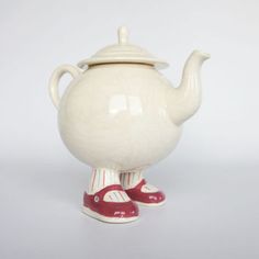 a white tea pot with red shoes on the bottom is shown in front of a gray background