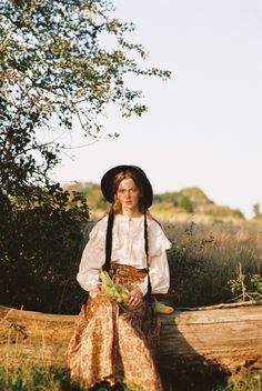 a woman sitting on top of a log wearing a hat and holding a plant in her hand