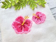 two pink flowers with green leaves on top of a white linen bag, sitting on a wooden surface