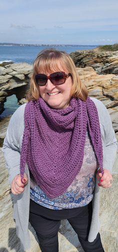 a woman is standing on some rocks wearing a purple crocheted scarf and sunglasses