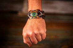 Embrace the fusion of craftsmanship and mystical allure with our Handmade Steampunk Bohemian Leather Cuff Bracelet. This unisex piece is a symphony of details, featuring an intricately carved and painted eyeball and mushrooms, surrounded by geometric patterns inspired by sacred geometry. The design is a bold testament to both artistic vision and spiritual symbolism. Rendered in rich, earthy tones of brown, green, and black, this bracelet interweaves multiple leather strips, creating a complex and textured aesthetic. The antique brass buckle closure adds a vintage touch while ensuring a comfortable fit for wrist sizes ranging from 7 to 9.25 inches. Perfect for festivals like Burning Man, spiritual gatherings, or everyday wear, this leather cuff serves as more than just an accessory—it's a p Hand Wrapped Spiritual Leather Bracelet For Festival, Spiritual Hand Wrapped Leather Bracelet For Festivals, Adjustable Steampunk Leather Bracelet, Adjustable Fantasy Jewelry For Festivals, Adjustable Fantasy Festival Jewelry, Unique Adjustable Leather Bangle Bracelet, Adjustable Unique Leather Bracelet For Festivals, Unique Hand Wrapped Leather Bracelet For Festivals, Handmade Adjustable Steampunk Bracelets