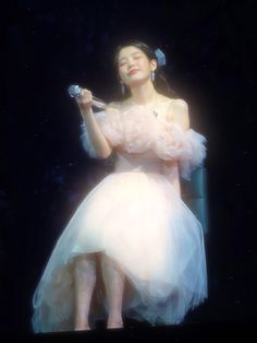 a woman in a white dress sitting on a chair holding a microphone and looking up at the sky