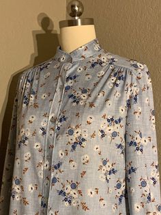 Absolutely darling 1970's button up blouse in a cute country floral print with crosshatch background, shades of blue, white and brown throughout. Blouse has blue pearlescent buttons up the front to a high collarless neckline. Beautiful ruching at the shoulders and long sleeves with single button cuffs. Made of a medium weight polyester fabric. Original brand tag no longer present, labeled a vintage size 11/12. Blouse would best fit a modern women's size medium (please see measurements below for Light Blue Buttoned Blouse For Fall, Classic Floral Print Tops For Daywear, Blue Blouse With Vintage Print For Spring, Light Blue Blouse For Fall Daywear, Vintage Blue Blouse For Fall, Spring Vintage Print Button-up Blouse, Vintage Spring Blouse With Covered Buttons, Vintage Light Blue Button-up Tops, Light Blue Vintage Button-up Tops