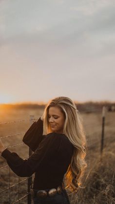 a woman standing next to a barbed wire fence with the sun setting in the background