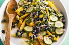 a white bowl filled with corn, cucumbers and black olives next to a wooden spoon