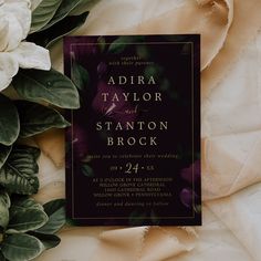 a wedding card with purple flowers and greenery on the table next to white roses